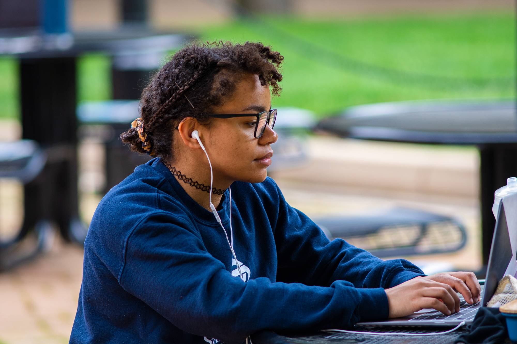 Student working on laptop outdoors with earbuds in ears.